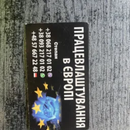 Czarna wizytówka z ukraińskim napisem 'Pracewłasztuwannia w Europi', numerami telefonów i grafiką dłoni na tle gwiazd Unii Europejskiej, leżąca na szarej, teksturowanej powierzchni.
