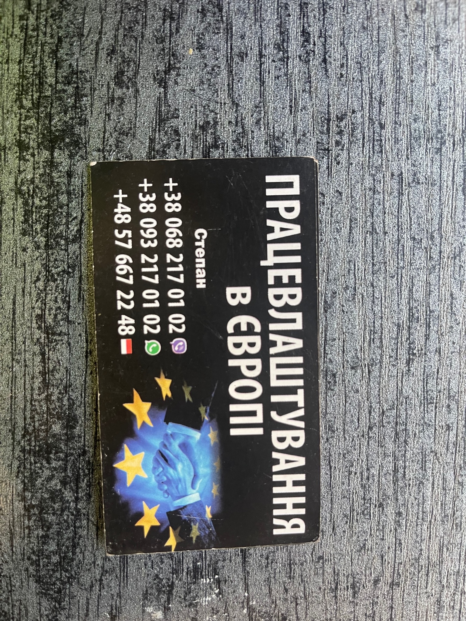 Czarna wizytówka z ukraińskim napisem 'Pracewłasztuwannia w Europi', numerami telefonów i grafiką dłoni na tle gwiazd Unii Europejskiej, leżąca na szarej, teksturowanej powierzchni.