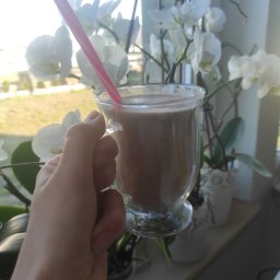 Zdrowejagodowe JAGODA DOMAGAŁA - Ręka trzyma szklankę z brązowym smoothie i różową słomką na tle okna z białymi orchideami.