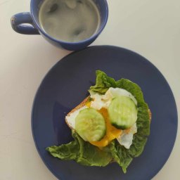 Zdrowejagodowe JAGODA DOMAGAŁA - Śniadanie na niebieskim talerzu: tost z jajkiem sadzonym, sałatą i plastrami ogórka, obok niebieski kubek z kawą.