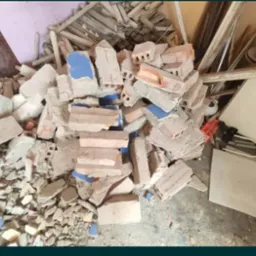 Stos gruzu i gruzowiska z fragmentami cegieł, płytek ceramicznych z niebieskimi elementami i resztkami drewna w pomieszczeniu po remoncie.