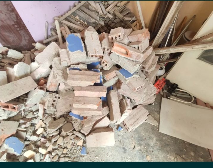 Stos gruzu i gruzowiska z fragmentami cegieł, płytek ceramicznych z niebieskimi elementami i resztkami drewna w pomieszczeniu po remoncie.