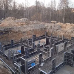 Szalunek fundamentów zbrojonych stalą, widok z góry na wykop z ziemią w tle, dwa niebieskie wiadra wewnątrz konstrukcji.