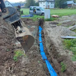 Głęboki wykop w ziemi z niebieską folią ochronną wzdłuż dna, łyżka koparki w tle, krajobraz wiejski z zabudowaniami w oddali.