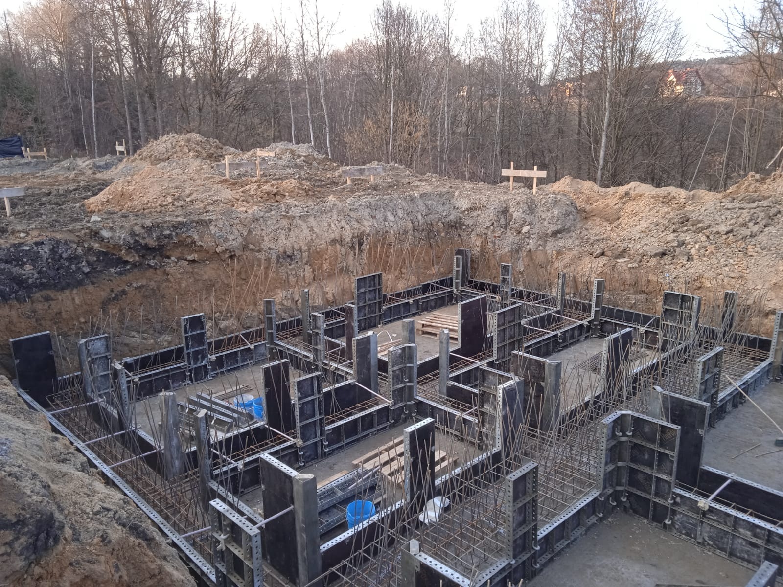 Szalunek fundamentów zbrojonych stalą, widok z góry na wykop z ziemią w tle, dwa niebieskie wiadra wewnątrz konstrukcji.