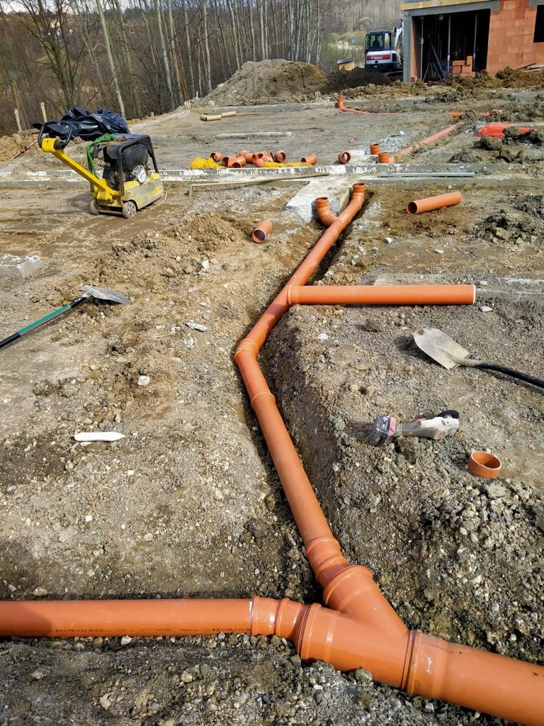 Pomarańczowe rury kanalizacyjne ułożone w wykopie na placu budowy, zagęszczarka w tle, widoczne narzędzia.