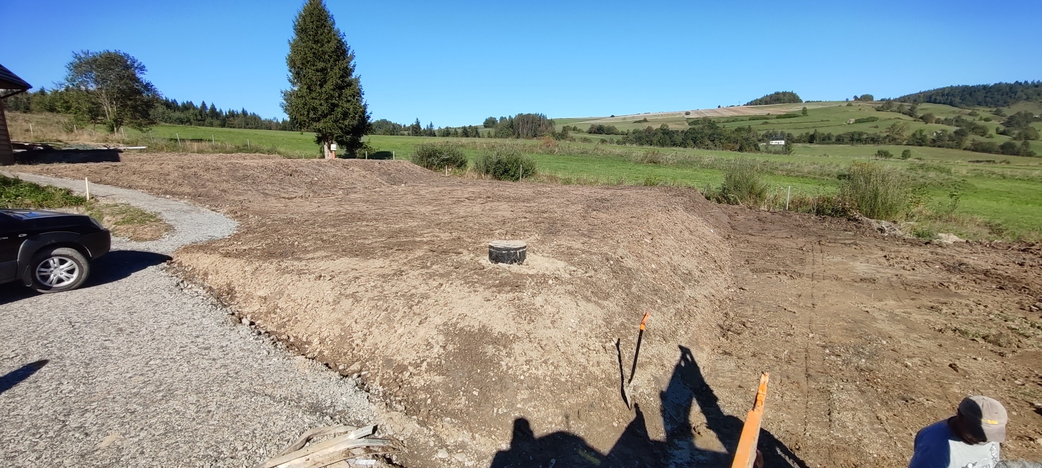 Teren po robotach ziemnych z widocznym włazem studzienki kanalizacyjnej na tle pagórkowatego krajobrazu i błękitnego nieba.