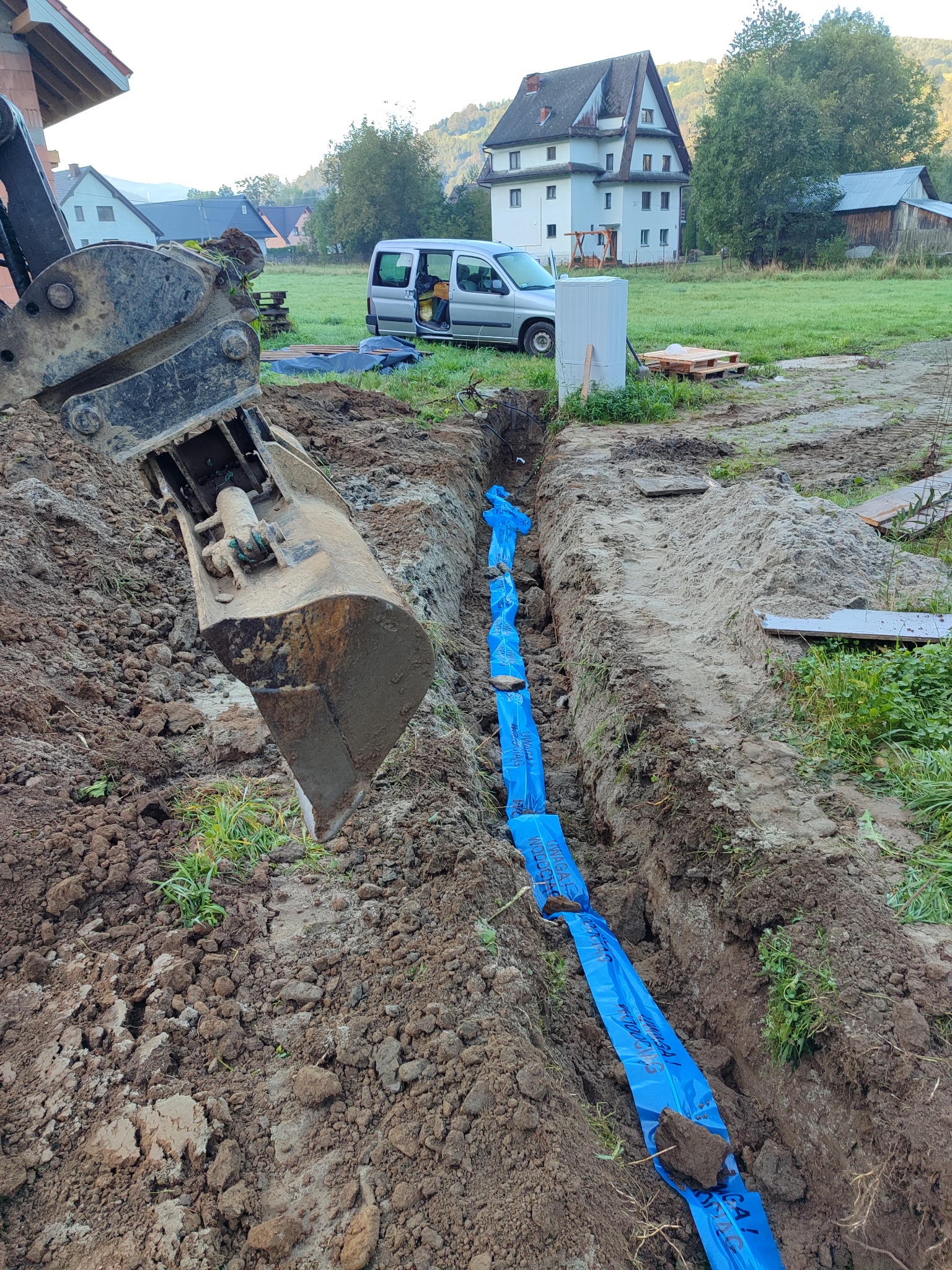 Głęboki wykop w ziemi z niebieską folią ochronną wzdłuż dna, łyżka koparki w tle, krajobraz wiejski z zabudowaniami w oddali.