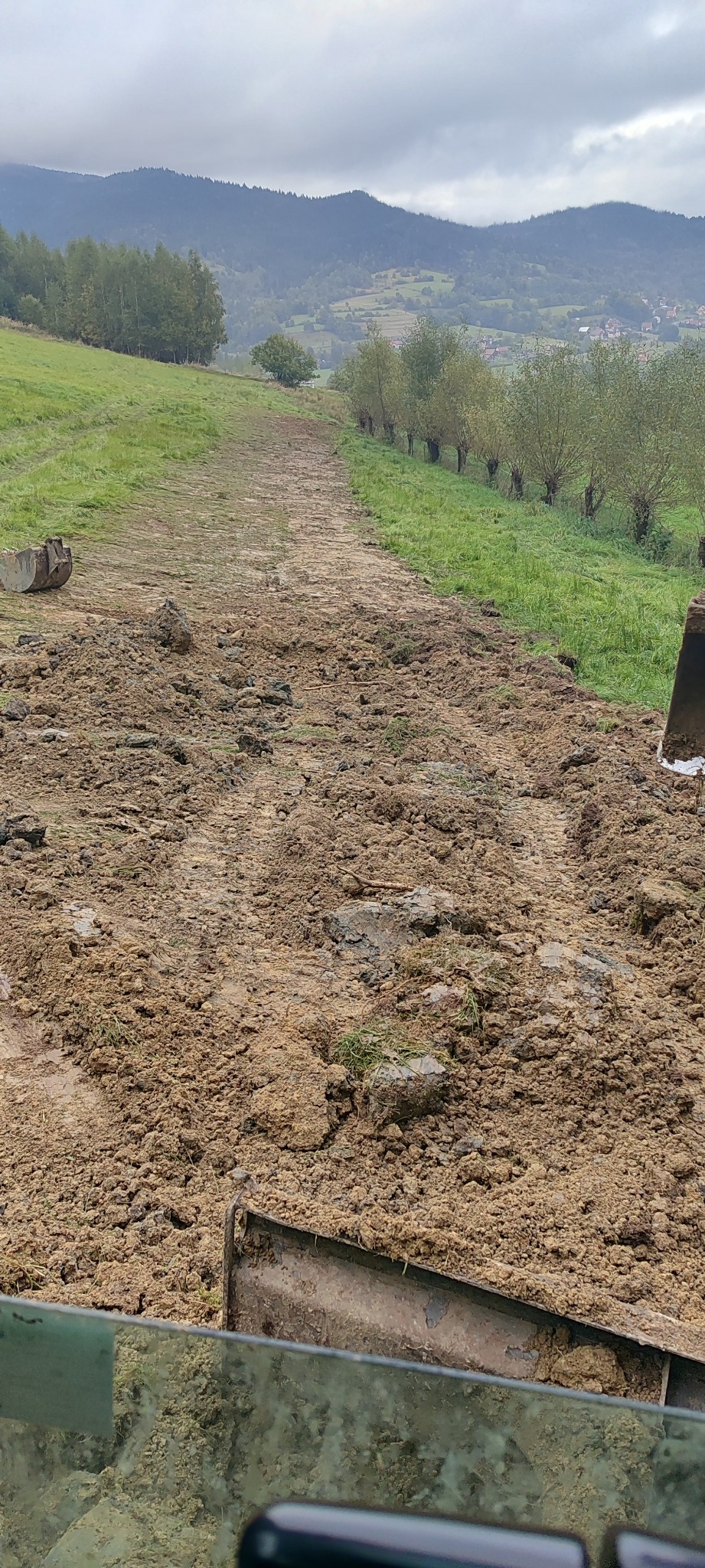 Świeżo przekopana ziemia z widocznymi śladami gąsienic koparki na tle krajobrazu z drzewami i wzgórzami w pochmurny dzień.