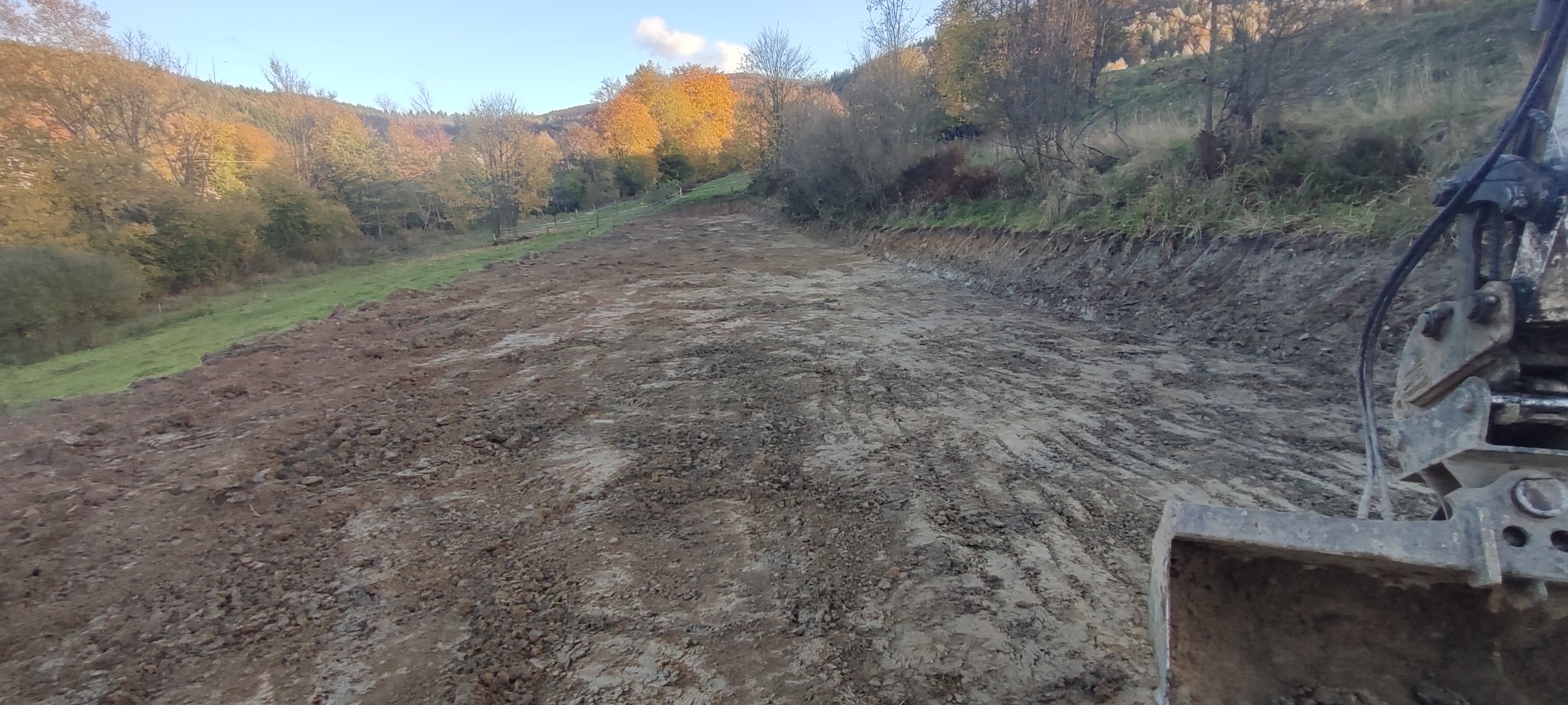 Przygotowany teren pod budowę, widok z kabiny koparki na wyrównaną, gliniastą powierzchnię z widocznymi śladami gąsienic, w tle zalesione wzgórza w jesiennej scenerii.