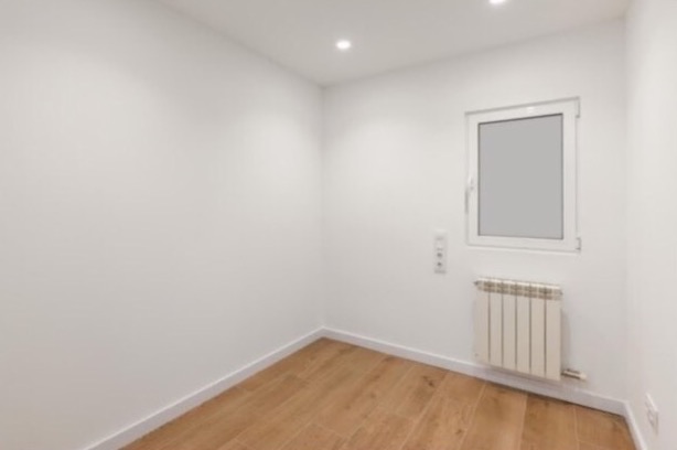 Pusty pokój po remoncie z białymi ścianami, drewnianą podłogą, grzejnikiem i oknem z białymi ramami.