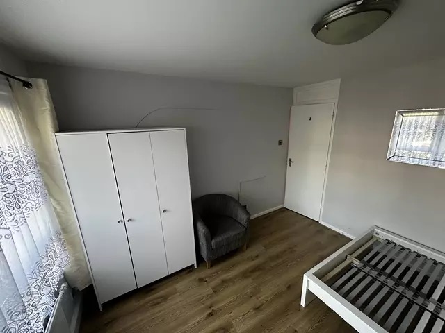 Pomalowane na szaro wnętrze pokoju z białym trzydrzwiowym ubraniowym, szarą tapicerowaną pufą, częściowo zmontowanym białym łóżkiem i białymi drzwiami.