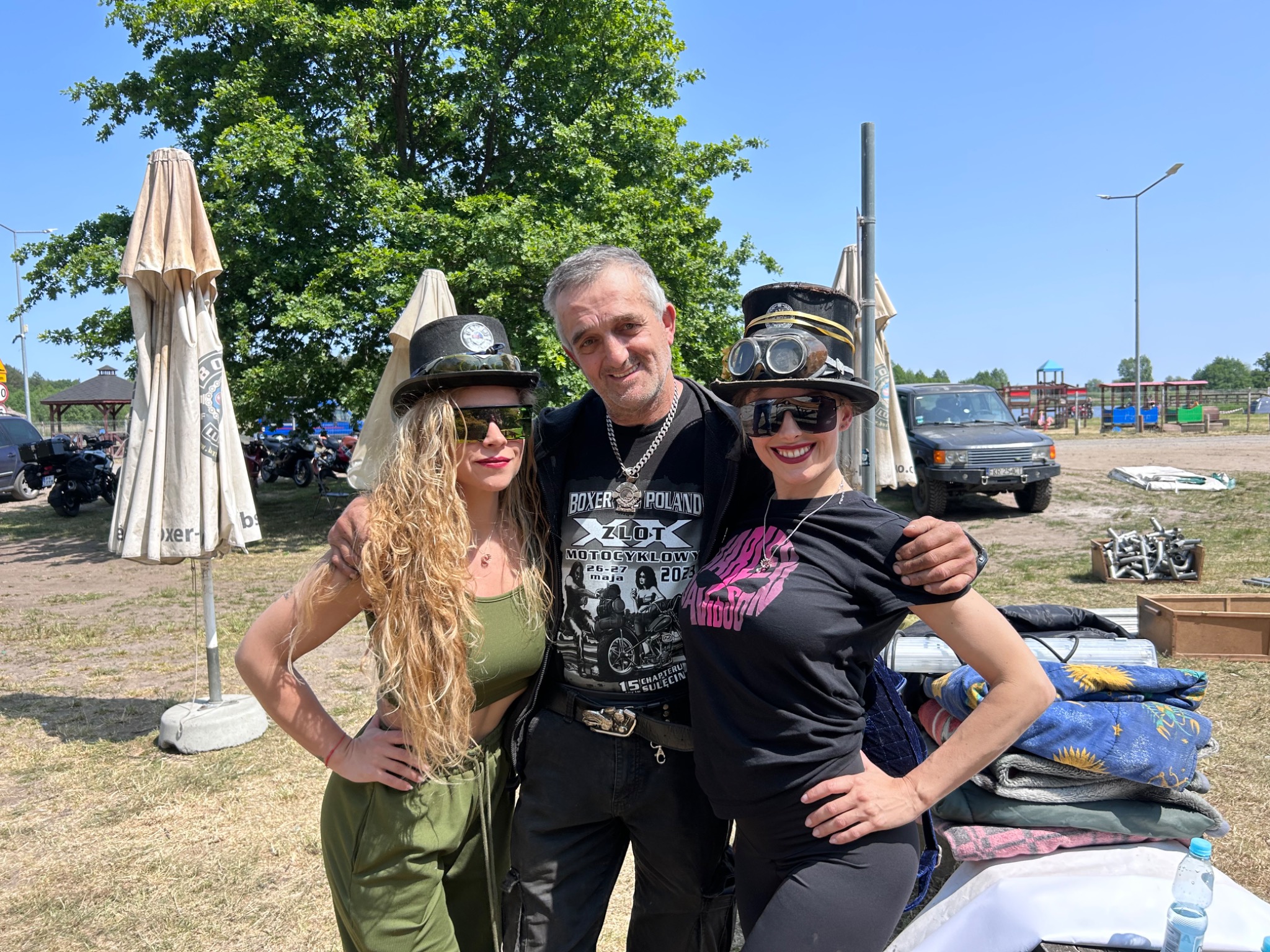 Mężczyzna obejmuje dwie kobiety w stylizowanych strojach na plenerowym wydarzeniu motocyklowym, w tle rozłożone parasole, motocykle i samochód terenowy.