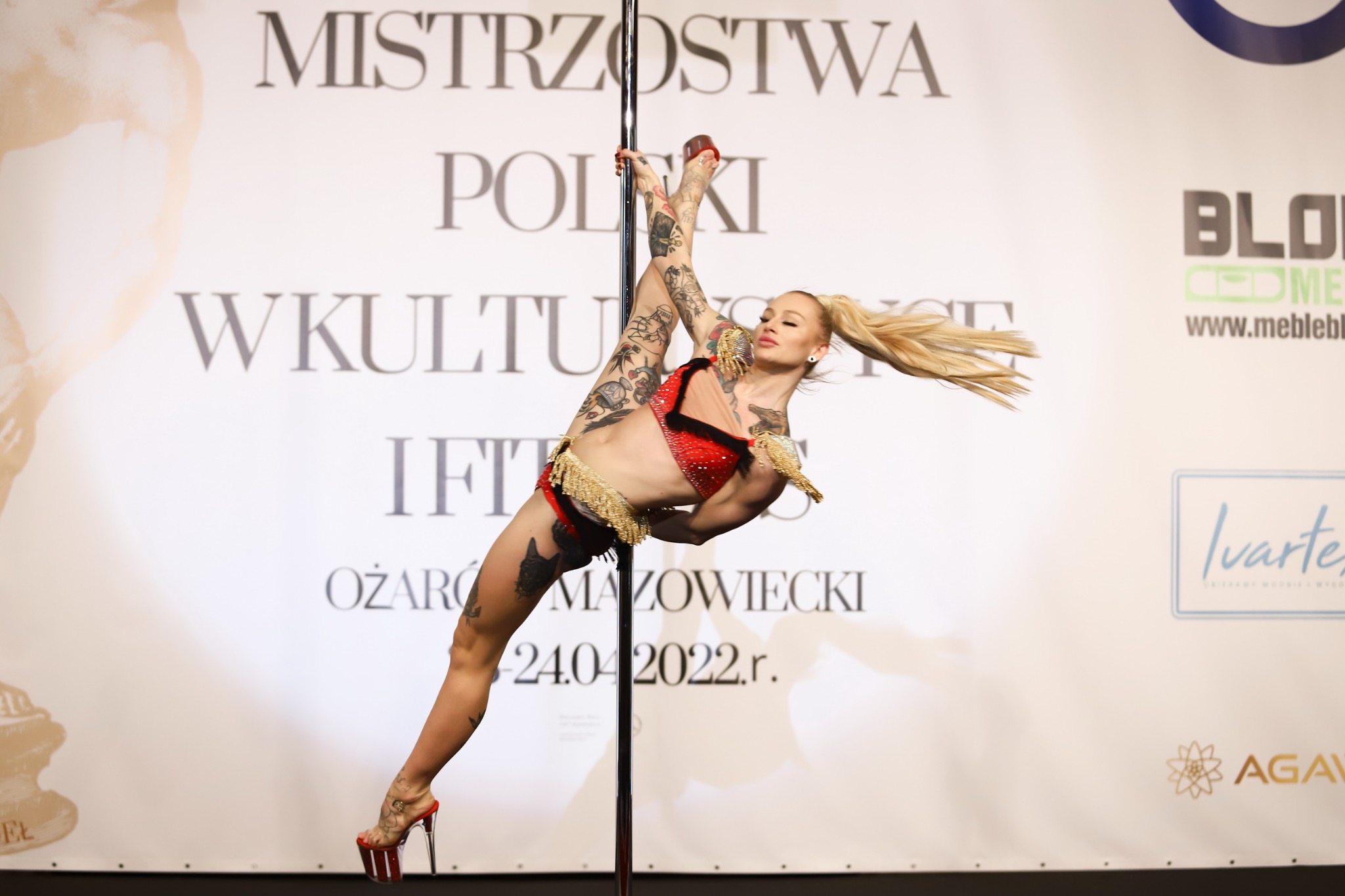 Kobieta z blond włosami i tatuażami w czerwonym stroju tanecznym wykonuje figurę na rurze podczas Mistrzostw Polski w Pole Dance w Ożarowie Mazowieckim 24.04.2022.