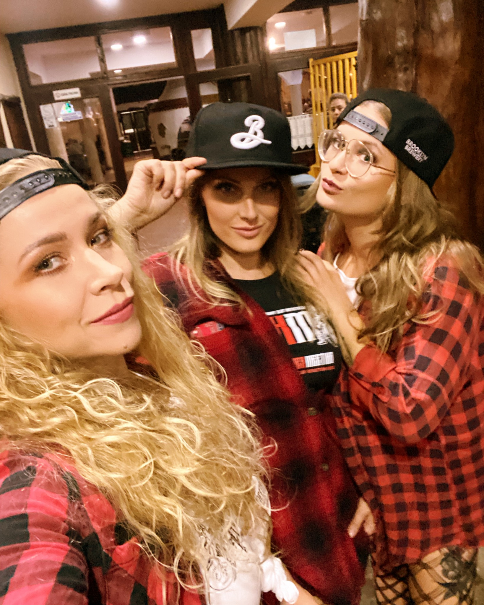 Trzy kobiety w czapkach i czerwonych koszulach w kratę pozują do selfie wewnątrz budynku z drewnianymi elementami wystroju.