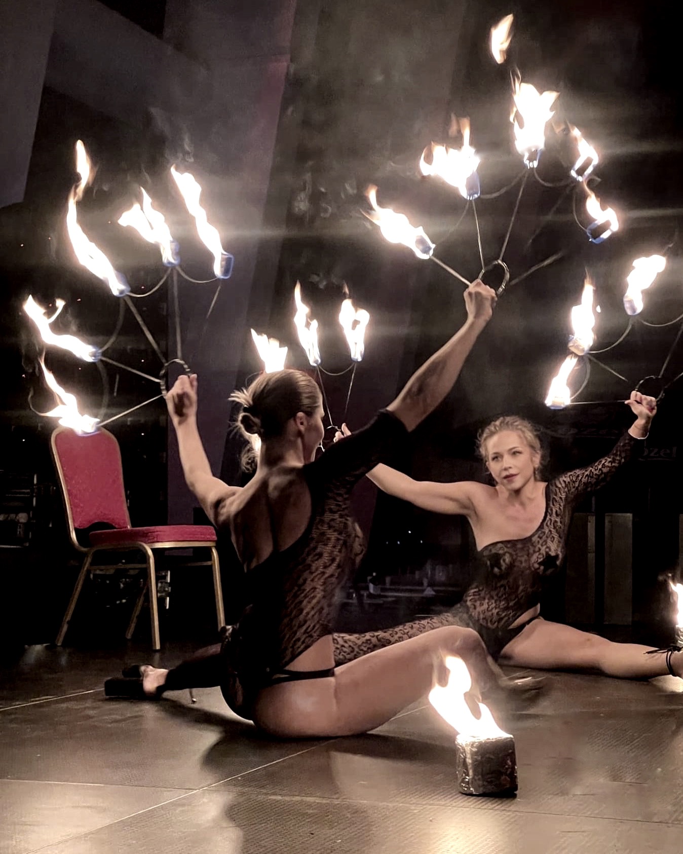 Dwie kobiety w czarnych strojach prezentują pokaz z ogniem na scenie, jedna trzyma płonące rekwizyty w dłoniach, druga ma płonący rekwizyt na podłodze obok niej, czerwone krzesło w tle.