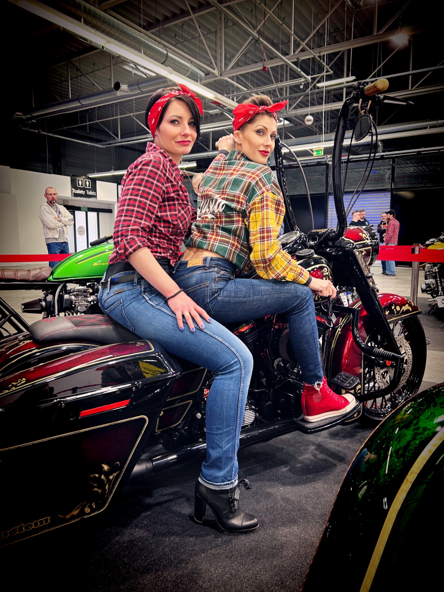 Dwie kobiety w stylizacjach retro pozują na motocyklu customowym, obie ubrane w kraciaste koszule i czerwone bandany, jedna siedzi z tyłu w dżinsach i czarnych botkach, druga siedzi z przodu...