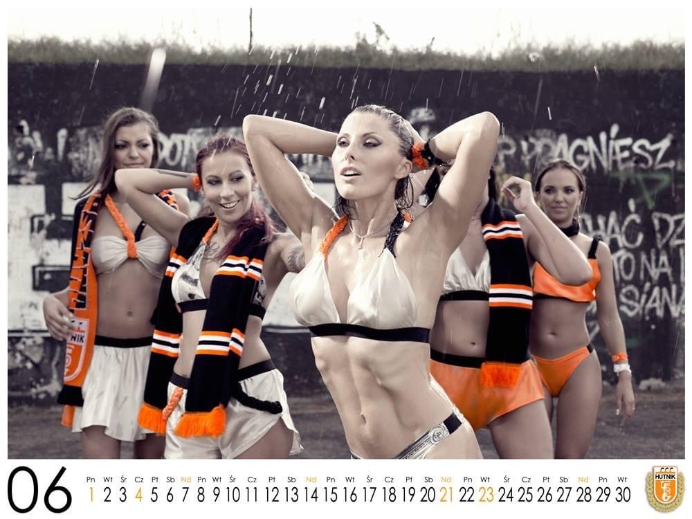 Grupa kobiet w sportowych strojach i szalikach w barwach pomarańczowo-czarnych pozuje w deszczu na tle graffiti, prawdopodobnie podczas sesji zdjęciowej.