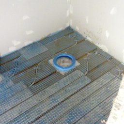 WOJCIECH WYSOCKI - Montaż ogrzewania podłogowego w łazience: siatka grzewcza z kablem ułożona na podłodze, widoczny otwór kanalizacyjny z niebieskim zabezpieczeniem. Ściany przygotowane do dalszych prac.