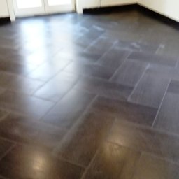 WOJCIECH WYSOCKI - Ułożona, ciemna podłoga z płytek w jodełkę, widok z niskiego kąta, z fragmentem ściany i drzwi balkonowych. Widoczne refleksy światła na powierzchni płytek.