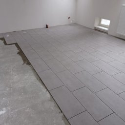 WOJCIECH WYSOCKI - Układanie szarych, prostokątnych płytek podłogowych w jasnym pomieszczeniu z oknem dachowym. Widoczne krzyżyki dystansowe i fragment surowej posadzki.