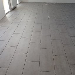 WOJCIECH WYSOCKI - Ułożone, szare płytki podłogowe w jasnym pomieszczeniu z białymi ścianami i grzejnikiem. Widoczne krzyżyki dystansowe między płytkami.