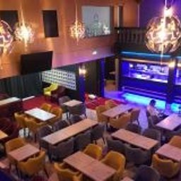 WOJCIECH WYSOCKI - Wnętrze restauracji z widokiem z góry: ciemne ściany, złote sufity, designerskie lampy wiszące, drewniane stoły z żółtymi i szarymi krzesłami, bar podświetlony niebieskim światłem w tle.