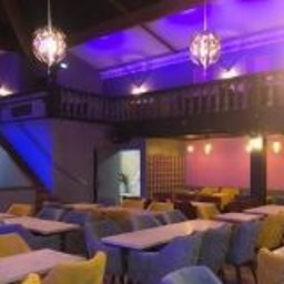 WOJCIECH WYSOCKI - Wnętrze restauracji z aranżacją stołów i krzeseł w odcieniach żółci i szarości, oświetlone dekoracyjnymi lampami i fioletowym światłem, widoczna antresola z ozdobną balustradą.