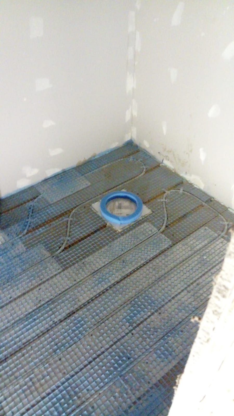 Montaż ogrzewania podłogowego w łazience: siatka grzewcza z kablem ułożona na podłodze, widoczny otwór kanalizacyjny z niebieskim zabezpieczeniem. Ściany przygotowane do dalszych prac.