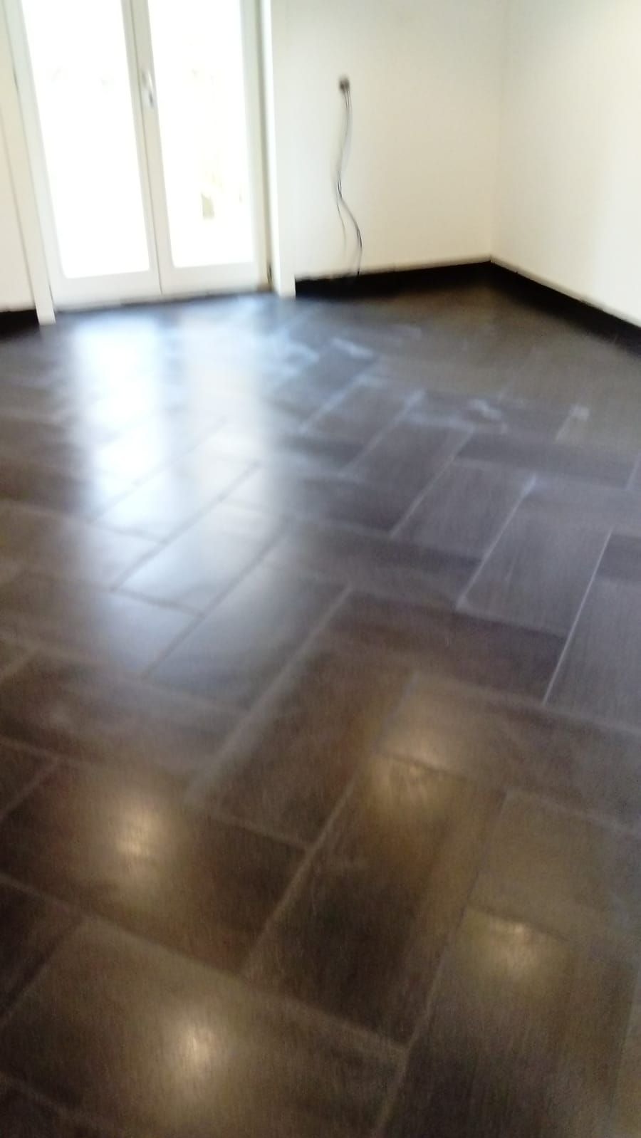 Ułożona, ciemna podłoga z płytek w jodełkę, widok z niskiego kąta, z fragmentem ściany i drzwi balkonowych. Widoczne refleksy światła na powierzchni płytek.