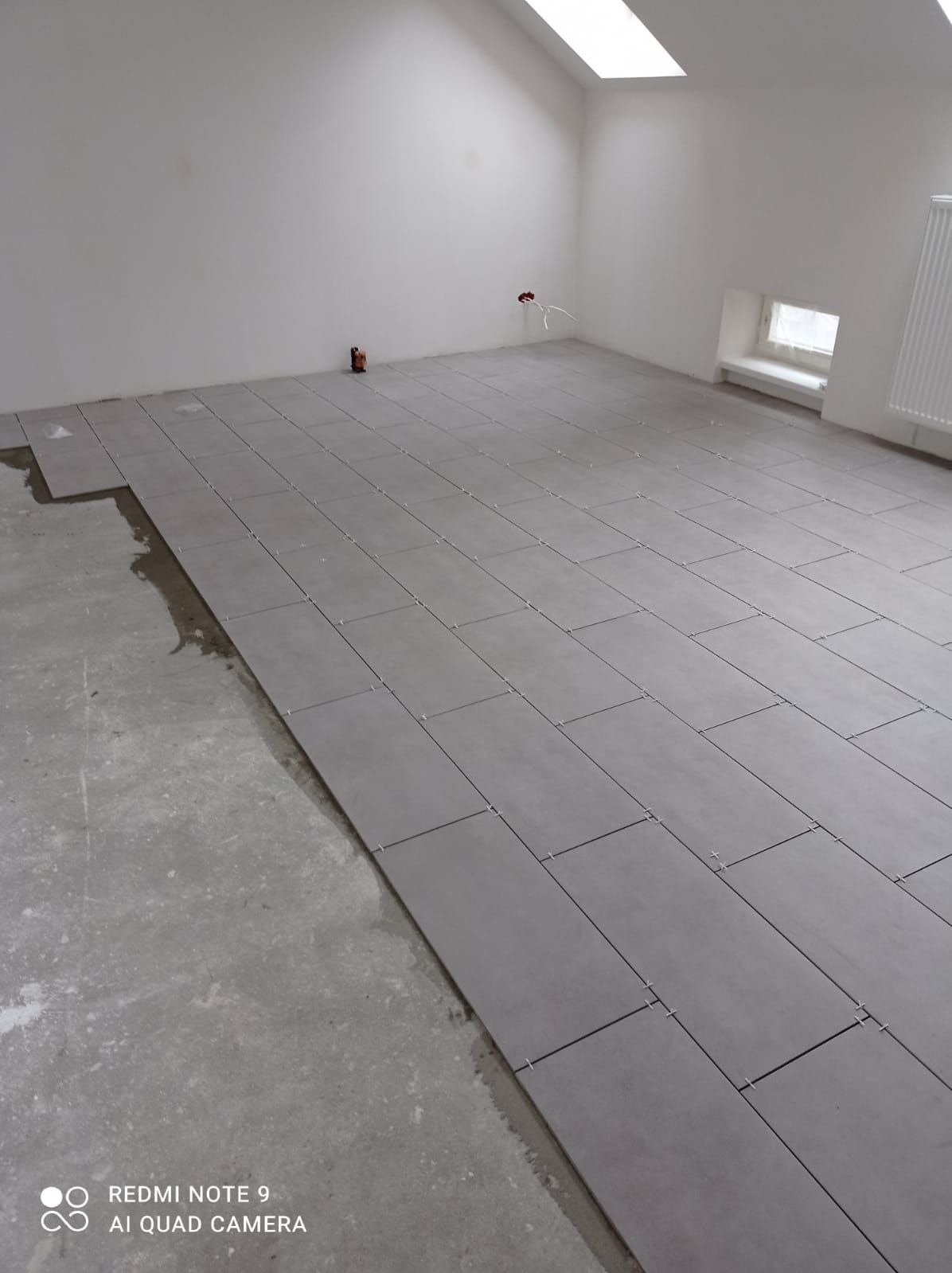 Układanie szarych, prostokątnych płytek podłogowych w jasnym pomieszczeniu z oknem dachowym. Widoczne krzyżyki dystansowe i fragment surowej posadzki.