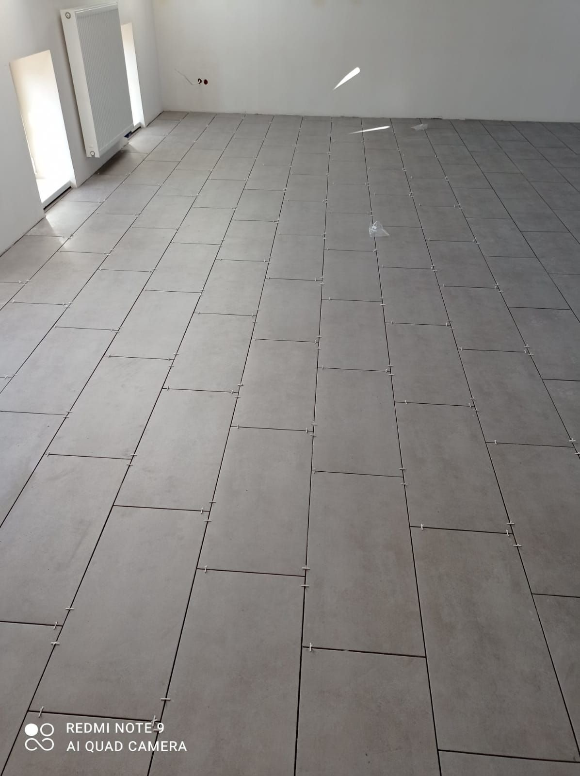 Ułożone, szare płytki podłogowe w jasnym pomieszczeniu z białymi ścianami i grzejnikiem. Widoczne krzyżyki dystansowe między płytkami.