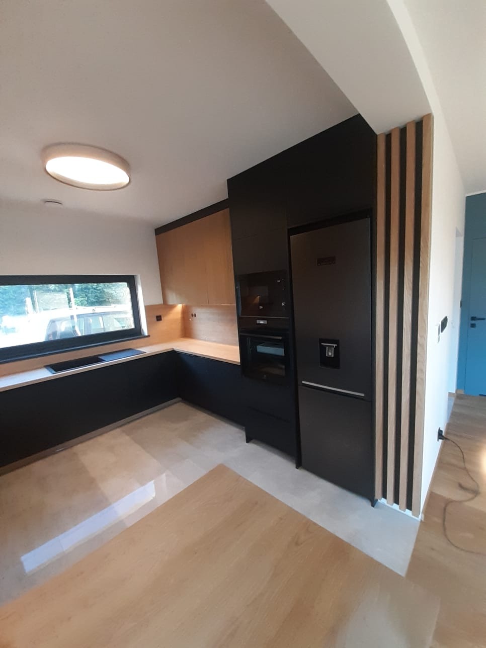 Nowoczesna kuchnia z czarnymi frontami i drewnianymi akcentami, oświetlona lampą sufitową, z widoczną lodówką i piekarnikiem w zabudowie.