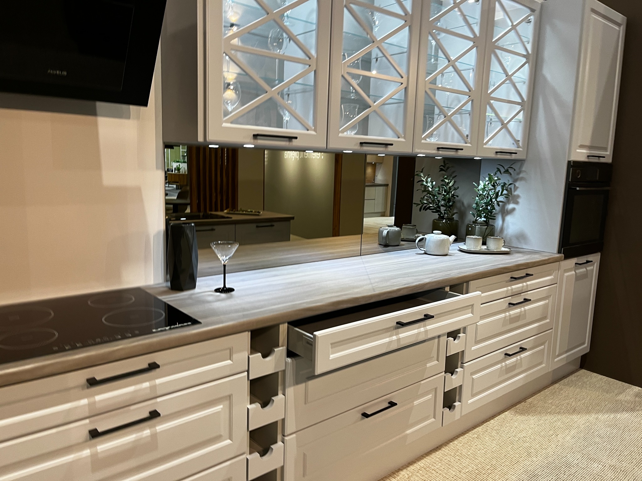 Biała zabudowa kuchenna z szarym blatem, otwartą szufladą i przeszklonymi szafkami górnymi z dekoracyjnymi krzyżami, odbijająca się w lustrzanym panelu nad blatem.