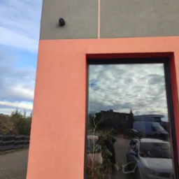 Kamera monitoringu i reflektor zamontowane na elewacji budynku, widok z zewnątrz, w oknie odbicie otoczenia.