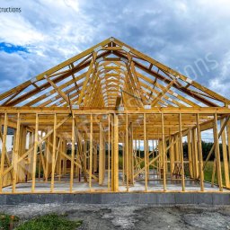 MT CONSTRUCTIONS - Wznoszenie drewnianej konstrukcji domu na betonowym fundamencie, widoczna więźba dachowa i elementy ścian, na zewnątrz trawa i błękitne niebo z chmurami.