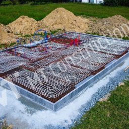 MT CONSTRUCTIONS - Przygotowany fundament pod dom z rozłożonym systemem ogrzewania podłogowego, widoczne zbrojenie i rury instalacji grzewczej, w tle sterta ziemi.