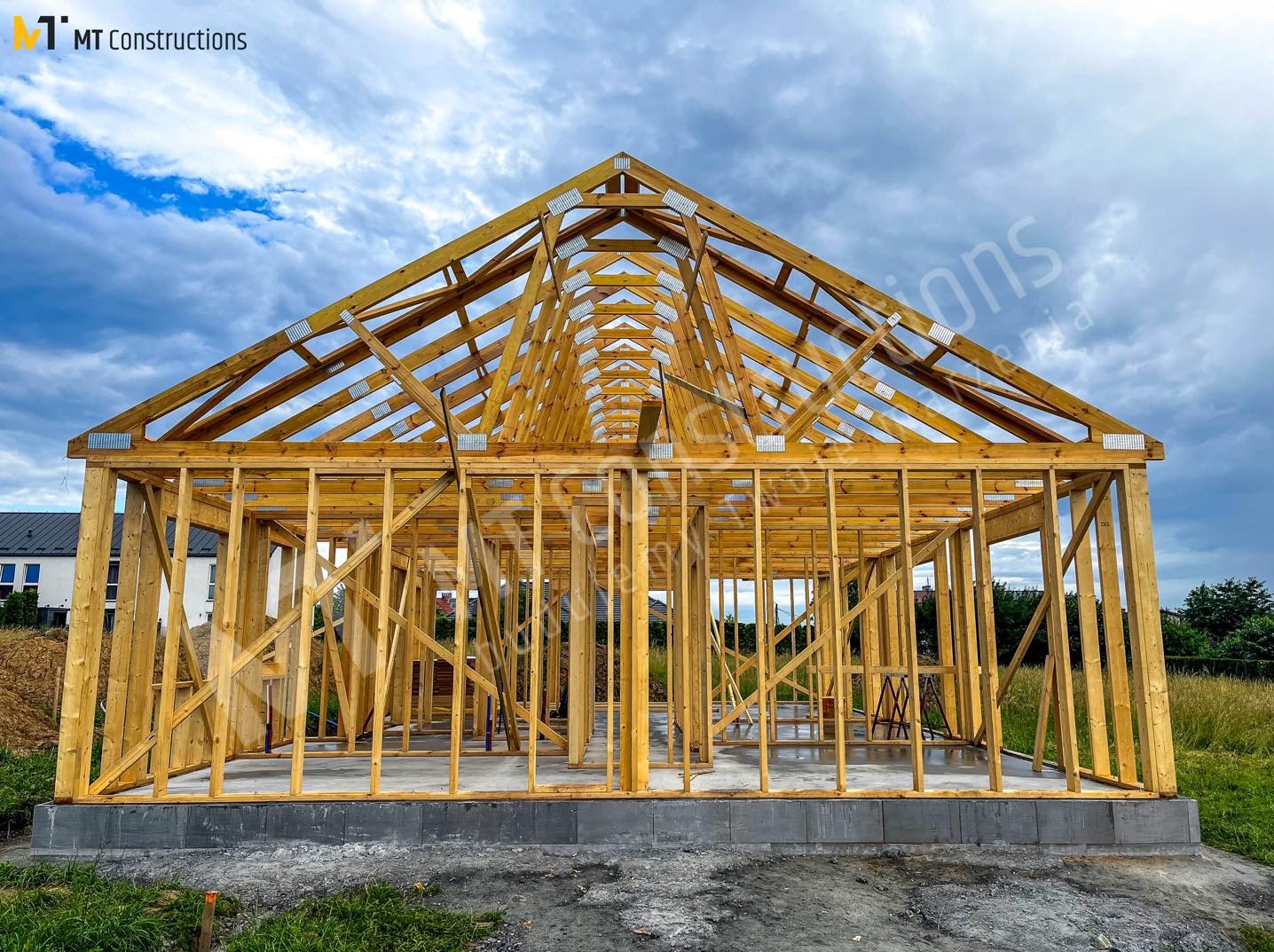 Wznoszenie drewnianej konstrukcji domu na betonowym fundamencie, widoczna więźba dachowa i elementy ścian, na zewnątrz trawa i błękitne niebo z chmurami.