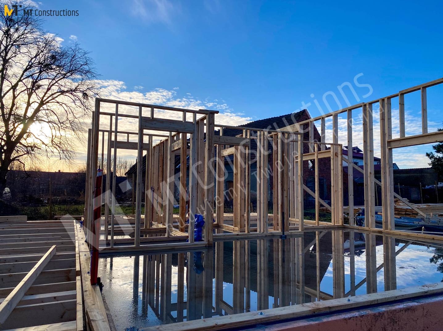 Wznoszenie drewnianej konstrukcji budynku na mokrym fundamencie, z refleksami nieba i drzew w wodzie, widoczne są elementy więźby dachowej i ściany szkieletowe.