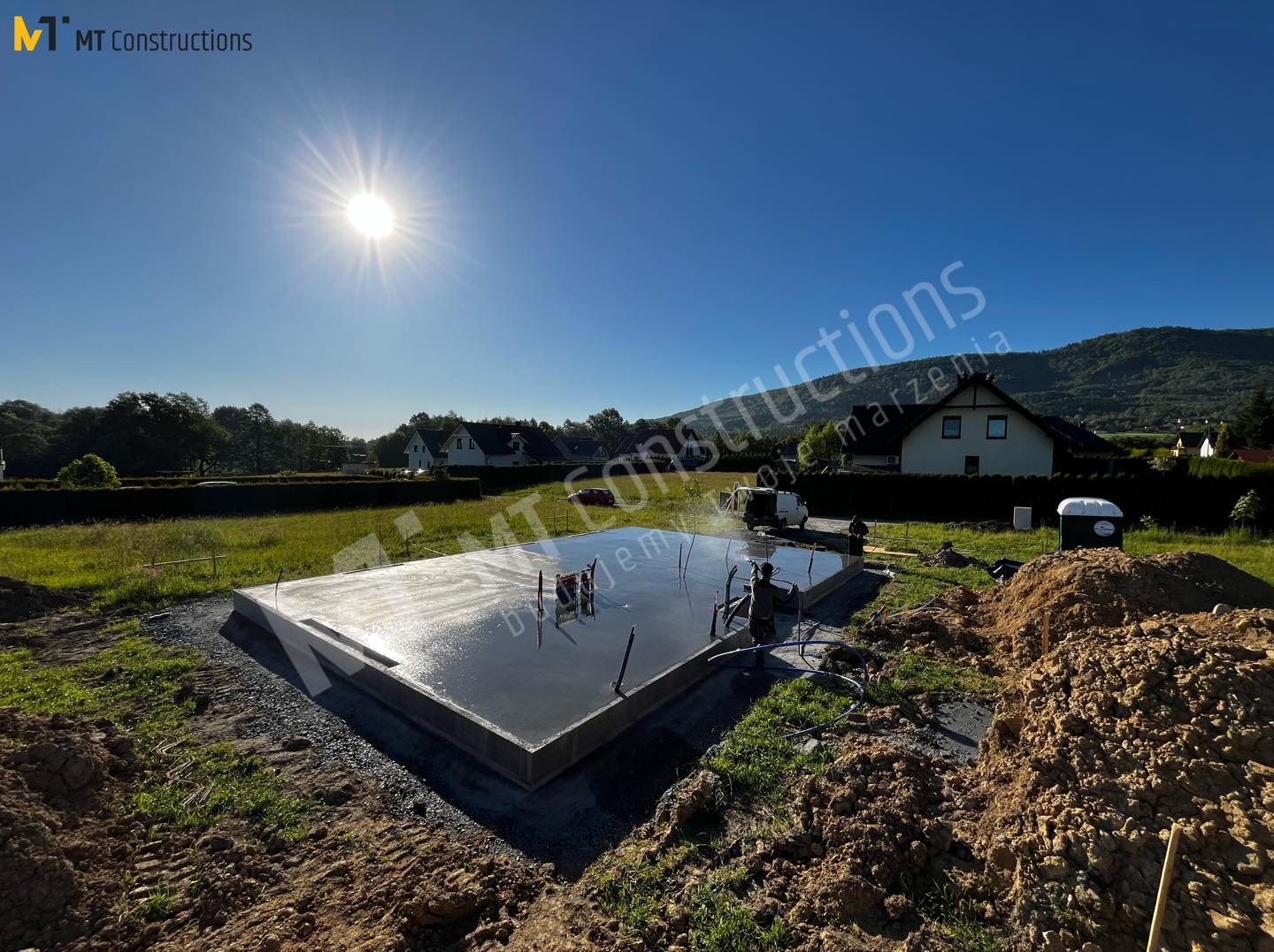 Świeżo wylana płyta fundamentowa z wystającymi prętami zbrojeniowymi, odbijająca promienie słoneczne, na tle zieleni i zabudowań jednorodzinnych pod błękitnym niebem.