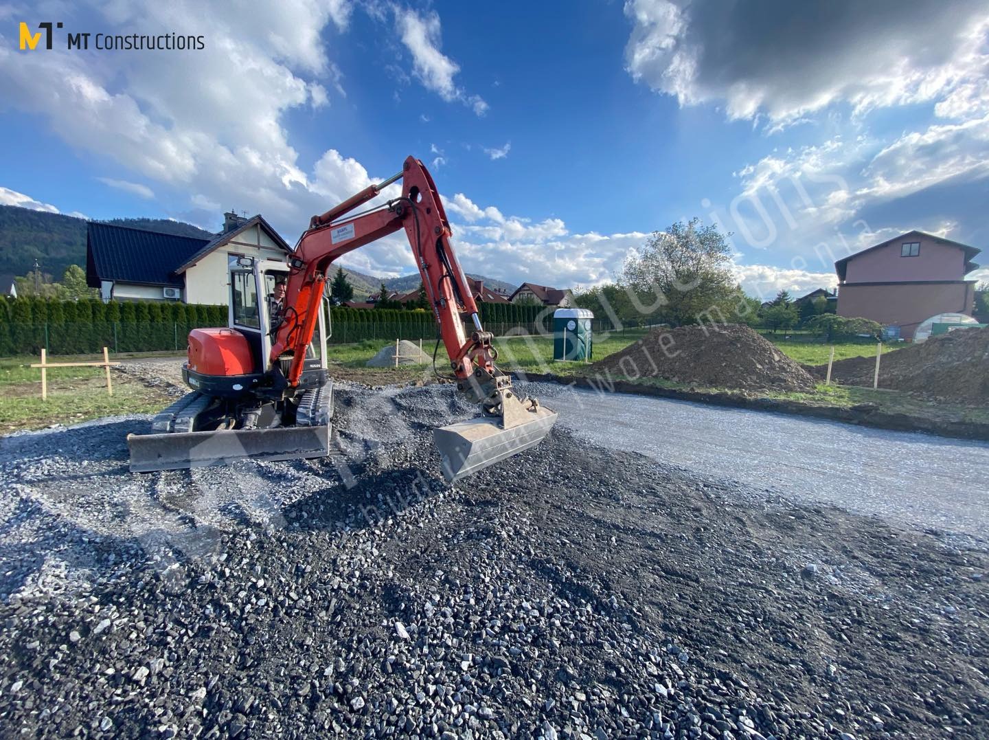 Czerwona mini koparka rozgarnia kamień na przygotowanym terenie budowy, widoczne fundamenty pod budynek w tle, błękitne niebo z chmurami.