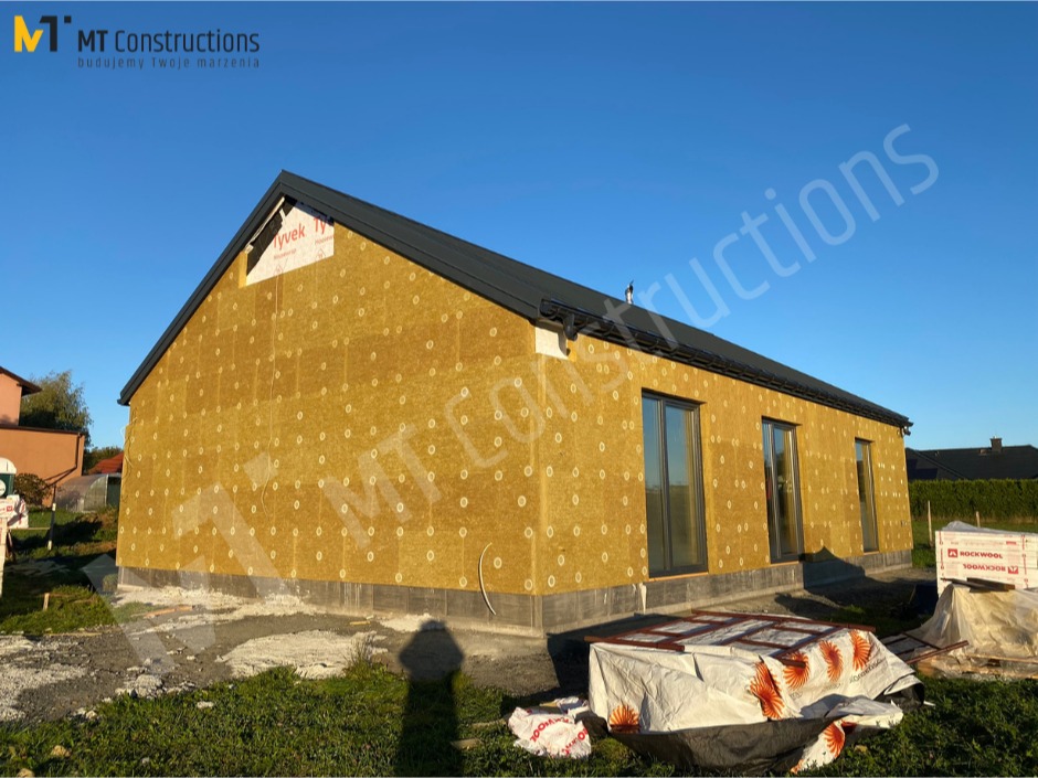 Budowa domu w trakcie ocieplania ścian zewnętrznych wełną mineralną, widoczne okna i czarny dach, na pierwszym planie materiały budowlane.