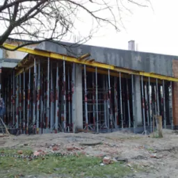 Wznoszenie ścian budynku z pustaków w Pile, widoczne liczne podpory szalunkowe i fragment zbrojenia stropu, w tle drzewo bez liści.