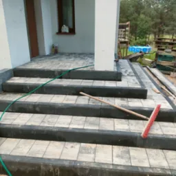 Wykończone schody zewnętrzne z kostki brukowej, z widocznym workiem z kruszywem, wężem ogrodowym i szczotką do zamiatania, otoczone zielenią.