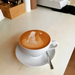 Cappuccino z latte art w kształcie łabędzia, podane w białej filiżance na spodku, obok łyżeczka, na jasnym blacie.