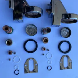 Rozłożone na białym tle chromowane elementy ekspresu do kawy, w tym dysze, uszczelki, sitka i śruby, widoczne ślady użytkowania na obudowach.