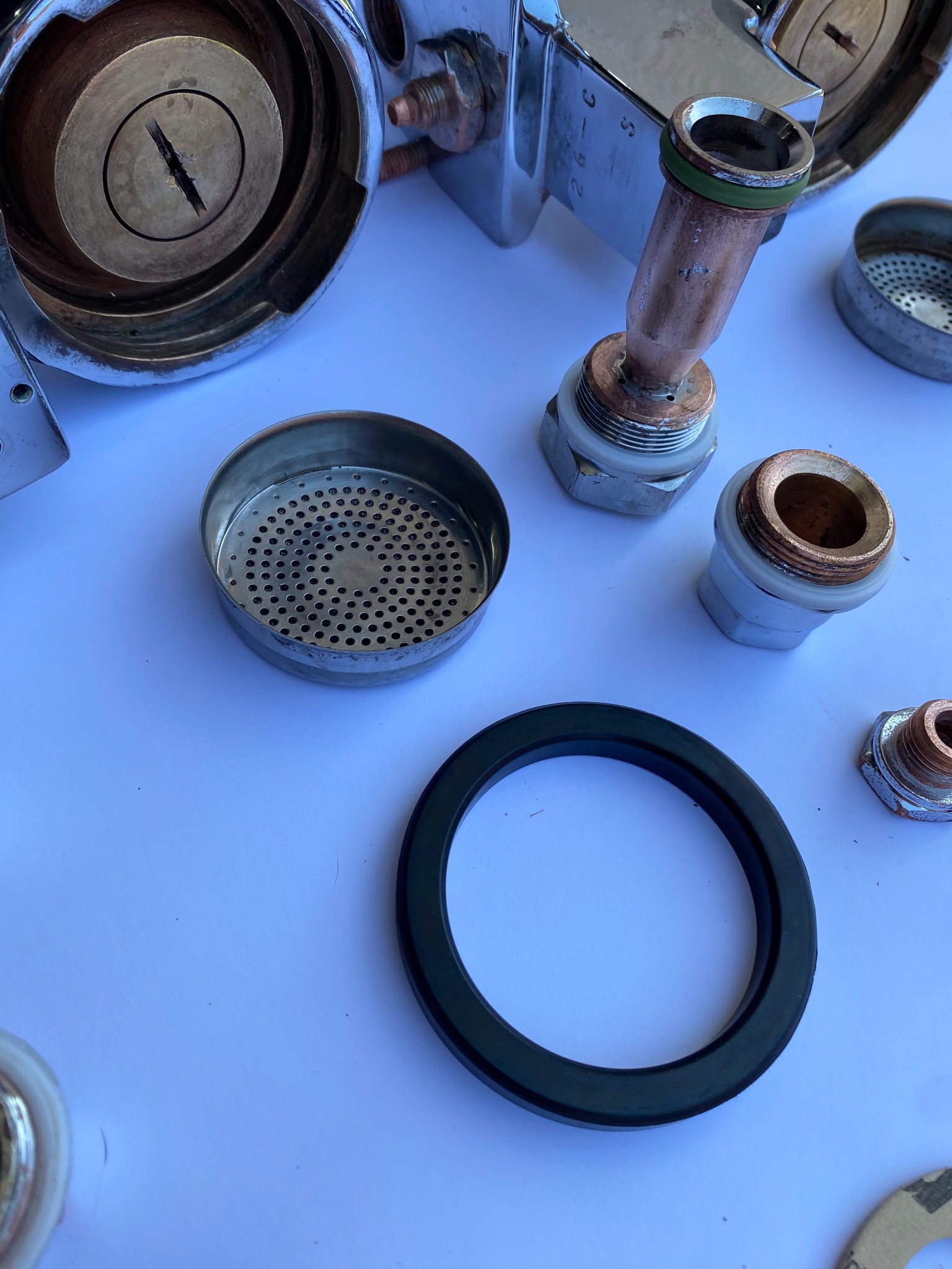 Rozłożone elementy ekspresu do kawy na białym tle: chromowana obudowa z mosiężnym wnętrzem, sitka, uszczelki i gwintowane złączki.