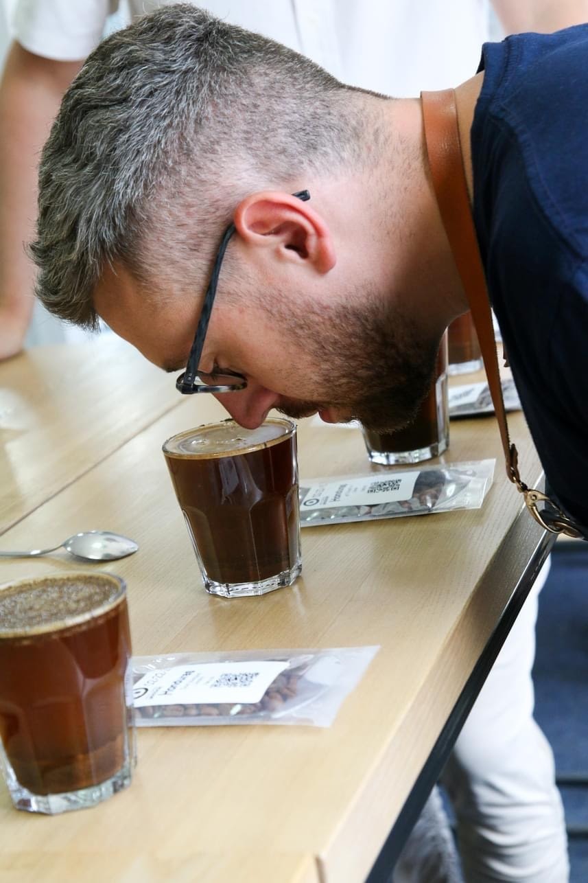 Mężczyzna z brodą w okularach pochyla się nad szklanką kawy, wąchając aromat. Na stole obok szklanki leżą ziarna kawy w przezroczystym opakowaniu i srebrna łyżeczka.