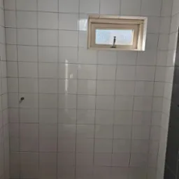 Łazienka w trakcie prac: białe, kwadratowe płytki na ścianach, małe okno z mleczną szybą, surowa podłoga przed położeniem płytek, widoczny odpływ liniowy w podłodze.