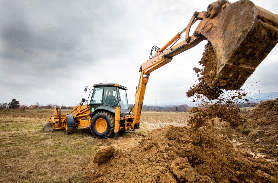 Żółta koparko-ładowarka na polu, wysypująca ziemię z łyżki, operator w kabinie, pochmurne niebo w tle.